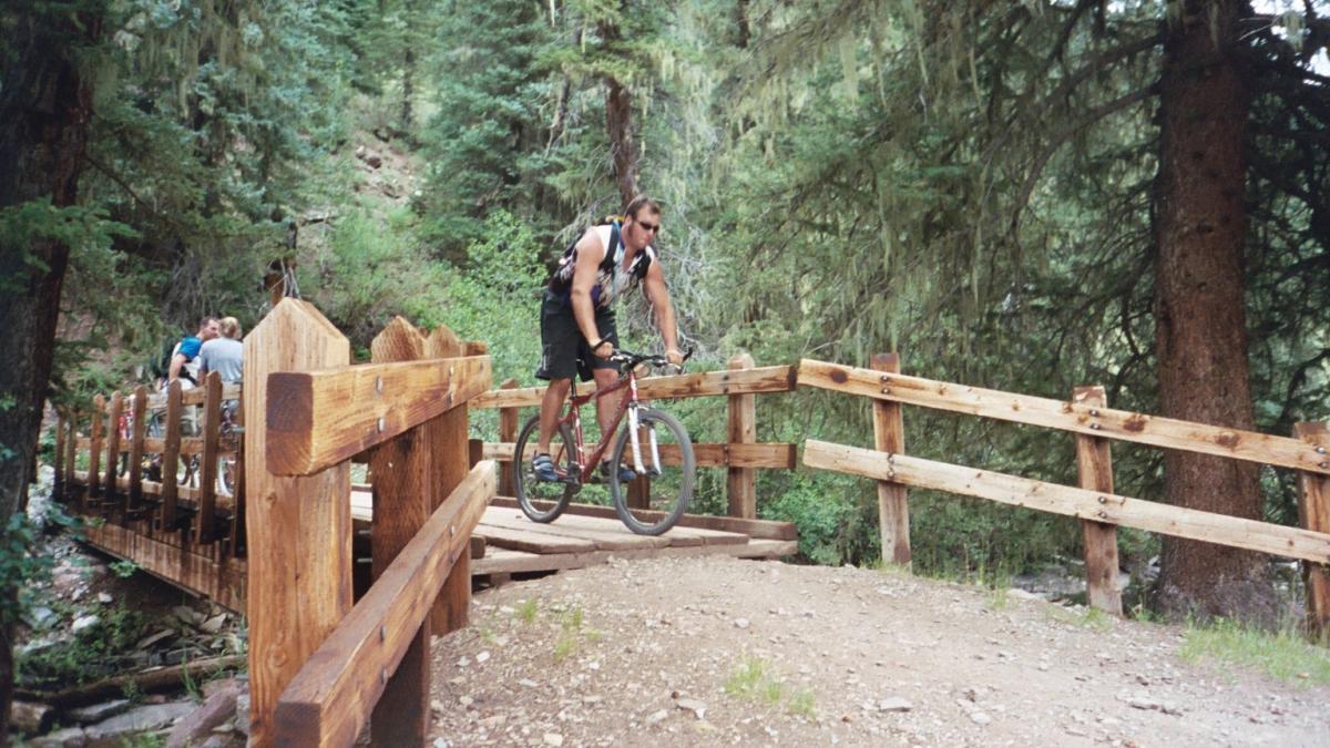 A mountain biker riding across a wooden bridge in a forested area, with trees surrounding the path. In the background, a few people are visible on the bridge. The scene captures an outdoor recreational setting. Hermosa Creek Trail mountain bike trail.