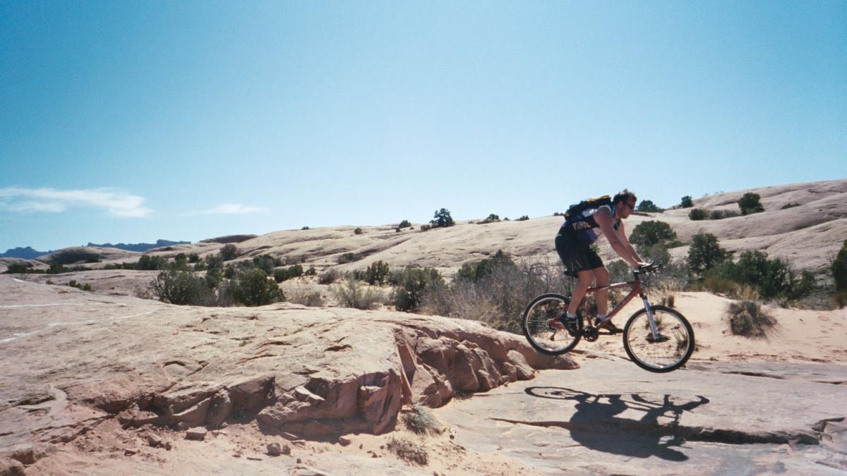 A mountain biker jumping over a rocky surface in a desert landscape with clear blue skies and distant hills in the background. Slickrock mountain bike trail.