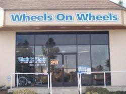 Storefront of a bicycle shop named "Wheels On Wheels," featuring a large sign above the entrance and a display of bicycles in the window. The shop has an "OPEN" sign visible and is situated in a well-lit area.