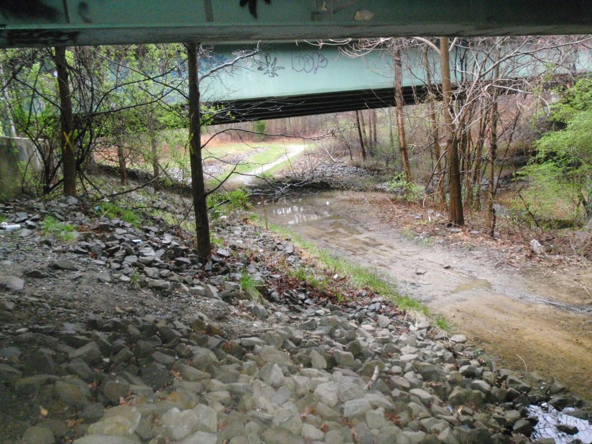 A view from beneath a green bridge, showcasing a rocky, uneven bank leading to a shallow stream. Sparse vegetation and bare trees are visible, with a dirt path meandering along the stream's edge. The setting has a natural yet slightly neglected appearance, with some litter scattered among the rocks. Cere's Park mountain bike trail.