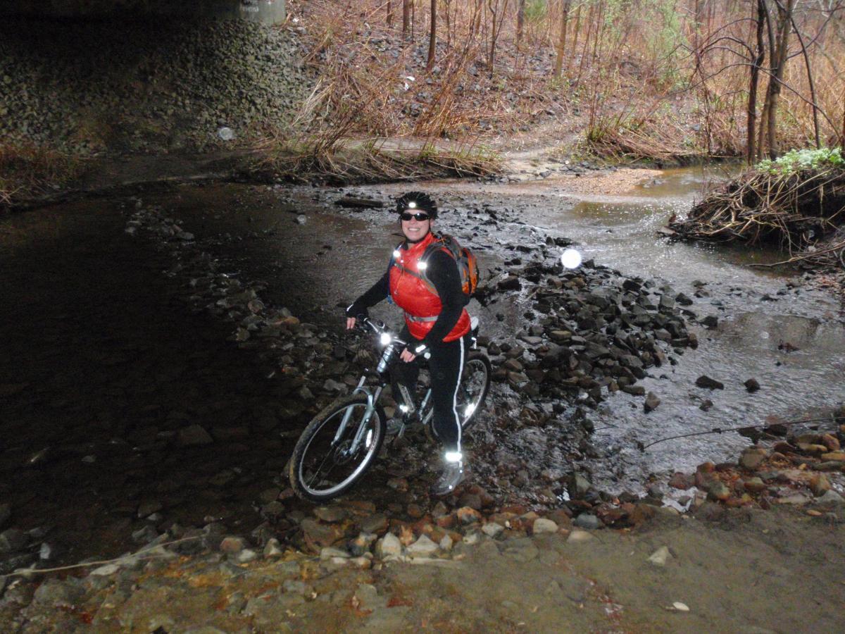 A person on a mountain bike, wearing a red vest and black pants, stands in shallow water surrounded by rocks under a bridge. The background features a forested area with sparse trees and no foliage, indicative of a rainy season. The scene conveys a sense of adventure in an outdoor environment. Cere's Park mountain bike trail.