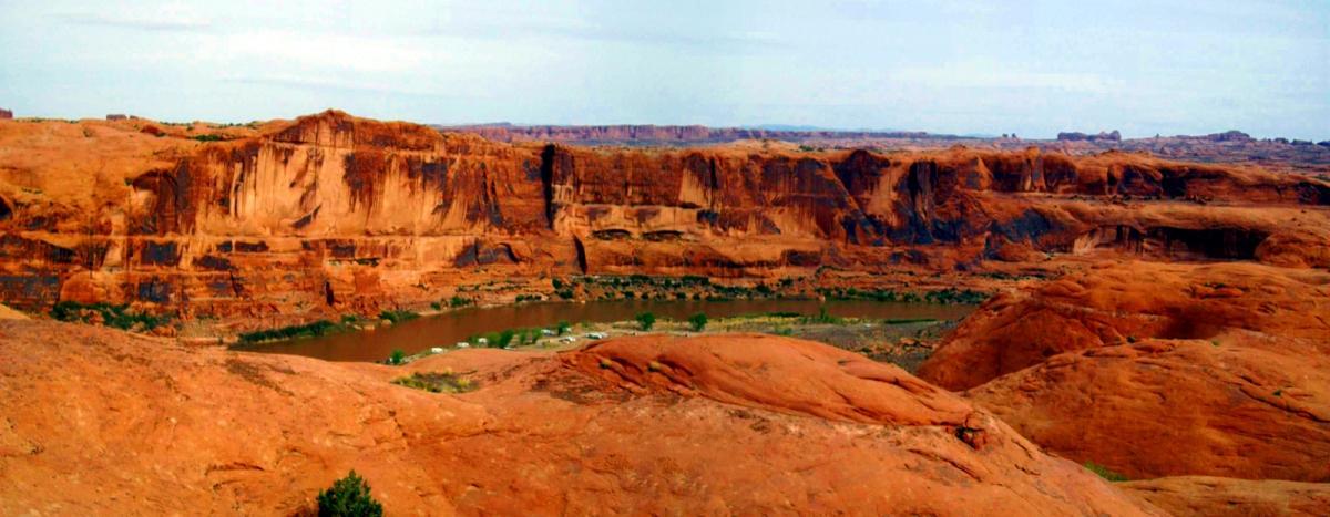 A panoramic view of a desert landscape featuring red rock formations and steep cliffs. A green river winds through the valley below, surrounded by patches of greenery and shrubs, under a clear blue sky. Slickrock mountain bike trail.
