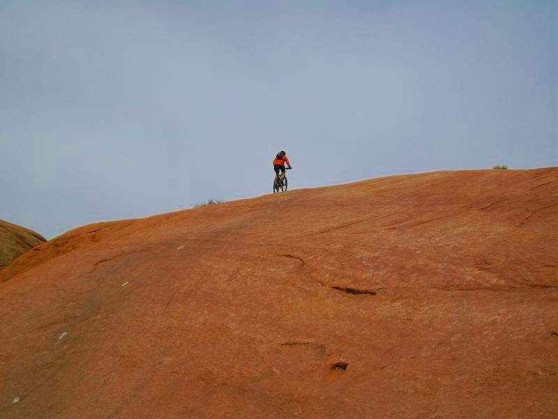 A person riding a mountain bike on a large, smooth, orange rock formation under a cloudy sky. Slickrock mountain bike trail.