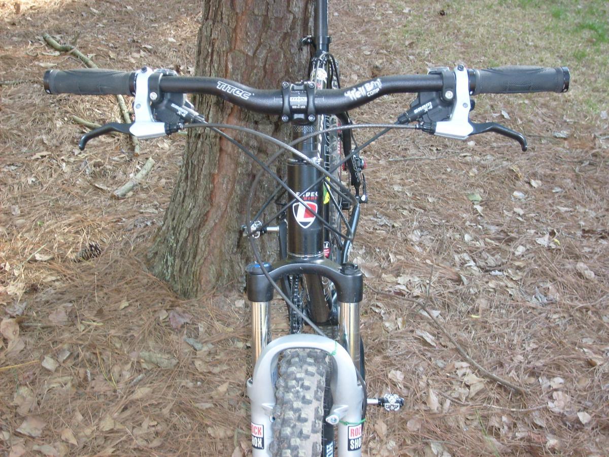 Mountain bike handlebars facing forward, positioned near a tree trunk, with pine needles and dried leaves on the ground. The bike features disc brakes and a suspension fork, showcasing a rugged design suitable for off-road riding. Tsali Right Loop mountain bike trail.