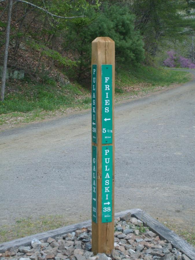 Wooden directional signpost indicating distances to Pulaski (38 miles), Fries (5.5 miles), and Galax (12 miles), set along a rural road in a forested area. New River Trail State Park mountain bike trail.