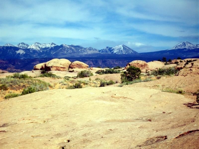 A rocky landscape featuring large, weathered boulders in the foreground, with sparse vegetation. In the background, a range of snow-capped mountains rises against a blue sky with scattered clouds. Slickrock mountain bike trail.