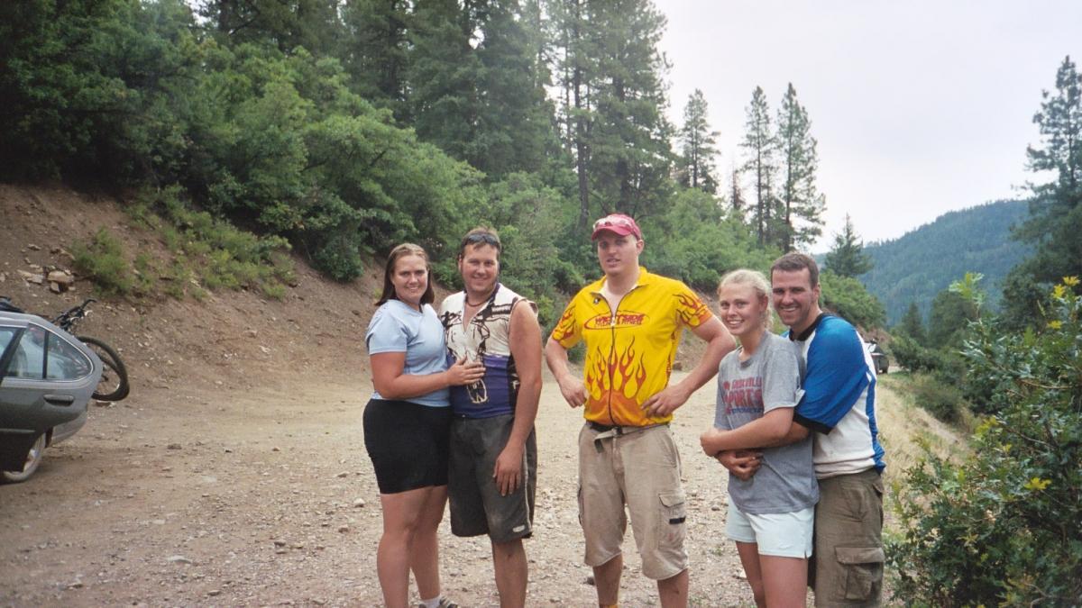 A group of five people stands together on a dirt road surrounded by trees and hills. They are smiling and posing for the camera, with two women and three men in casual outdoor clothing. A car and bicycles are visible in the background, suggesting a day of outdoor activities. The atmosphere is cheerful and friendly. Hermosa Creek Trail mountain bike trail.