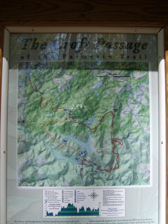 Map of the passage of the Palmetto Trail showing trails, points of interest, and elevation profiles. Features include marked routes in red and blue, various symbols for amenities, and a compass rose in the corner. The map is displayed in a wooden frame with surrounding greenery visible. Croft Passage mountain bike trail.