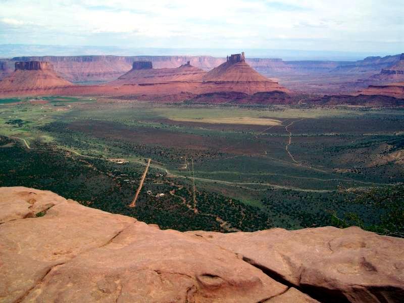 A panoramic view of a vast desert landscape featuring towering red rock formations and plateaus, with a green valley and winding dirt roads in the foreground beneath a cloudy sky. Porcupine Rim mountain bike trail.
