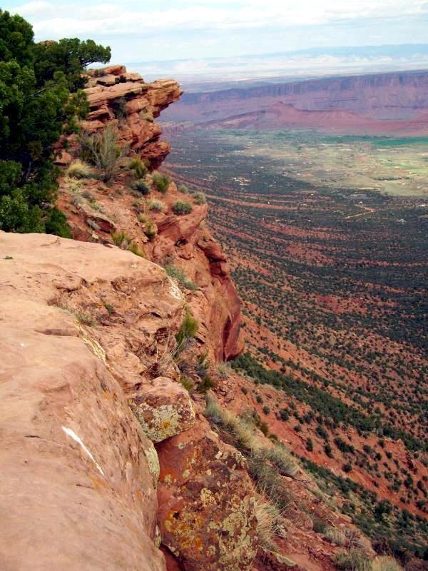 A panoramic view from the edge of a rocky cliff, showcasing a vast landscape of red rock formations, green vegetation, and distant mountains under a cloudy sky. Porcupine Rim mountain bike trail.