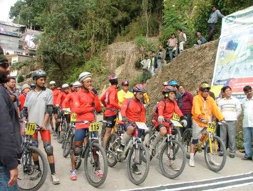 A group of cyclists in colorful jerseys and helmets prepare for a race, lined up on a road with spectators in the background. The cyclists include both adults and youths, and various bicycles are visible. The scene is set in a hilly area, with greenery and a banner promoting the event nearby.