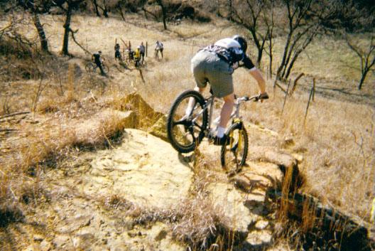A cyclist in a helmet rides down a rocky slope, navigating through dry grass and sparse trees. In the background, a group of bikers can be seen observing the descent. The scene captures the thrill of mountain biking in a natural terrain. Kanopolis Lake mountain bike trail.