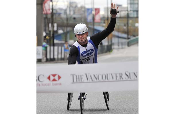 A wheelchair athlete raises one hand in victory as they cross the finish line, breaking the tape at a race. The setting includes urban scenery in the background, indicating a lively event atmosphere. The athlete is wearing a helmet and racing attire, showcasing determination and achievement.