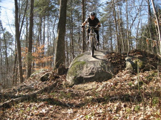 A mountain biker in a helmet is jumping off a large rock while riding on a forest trail with trees in the background. Fallen leaves and rocks are scattered on the ground, indicating a rugged terrain. Salem Lake mountain bike trail.