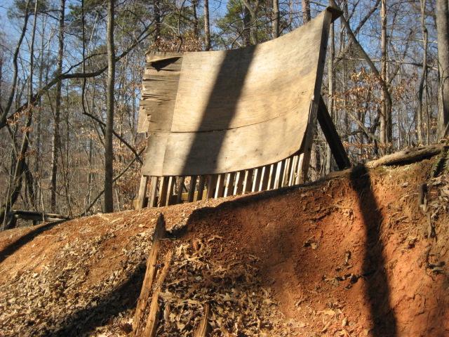 An inclined wooden ramp leaning against a dirt hillside, surrounded by trees and fallen leaves in a forested area. Salem Lake mountain bike trail.