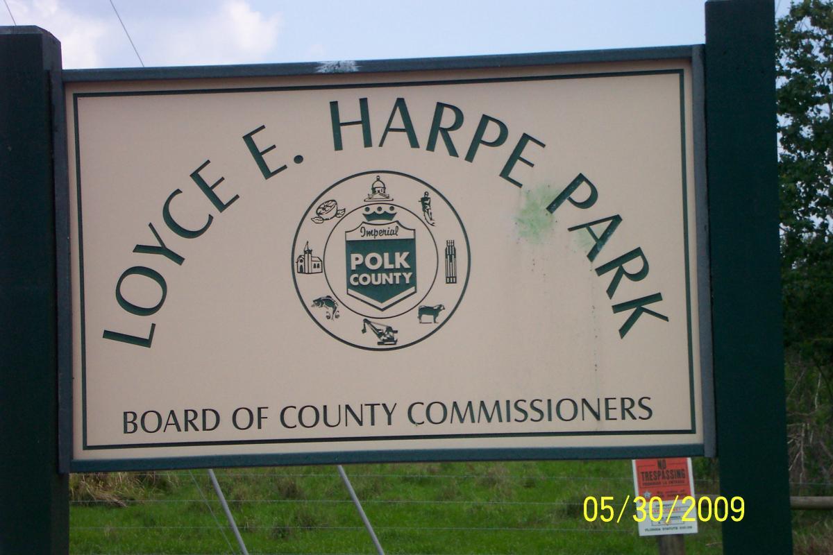 A park sign for Loyce E. Harpe Park, displaying the name prominently at the top, along with the seal of Polk County. The sign is framed in green with a light-colored background, and it includes the text "BOARD OF COUNTY COMMISSIONERS" at the bottom. A "No Trespassing" sign is visible in the background. The date on the bottom reads 05/30/2009. Loyce E. Harpe Park mountain bike trail.