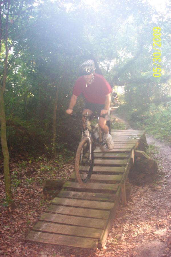 A person riding a mountain bike over a wooden bridge on a forest trail. The scene is surrounded by lush greenery, and the path is slightly muddy, indicating recent rain. The rider is wearing a helmet and a red shirt. Loyce E. Harpe Park mountain bike trail.