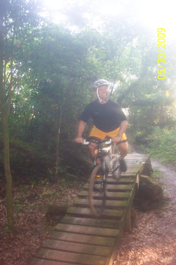 A person riding a mountain bike on a wooden bridge in a forested area, wearing a black shirt and yellow shorts, with a helmet on. The scene is slightly hazy, indicating bright sunlight filtering through the trees. Loyce E. Harpe Park mountain bike trail.