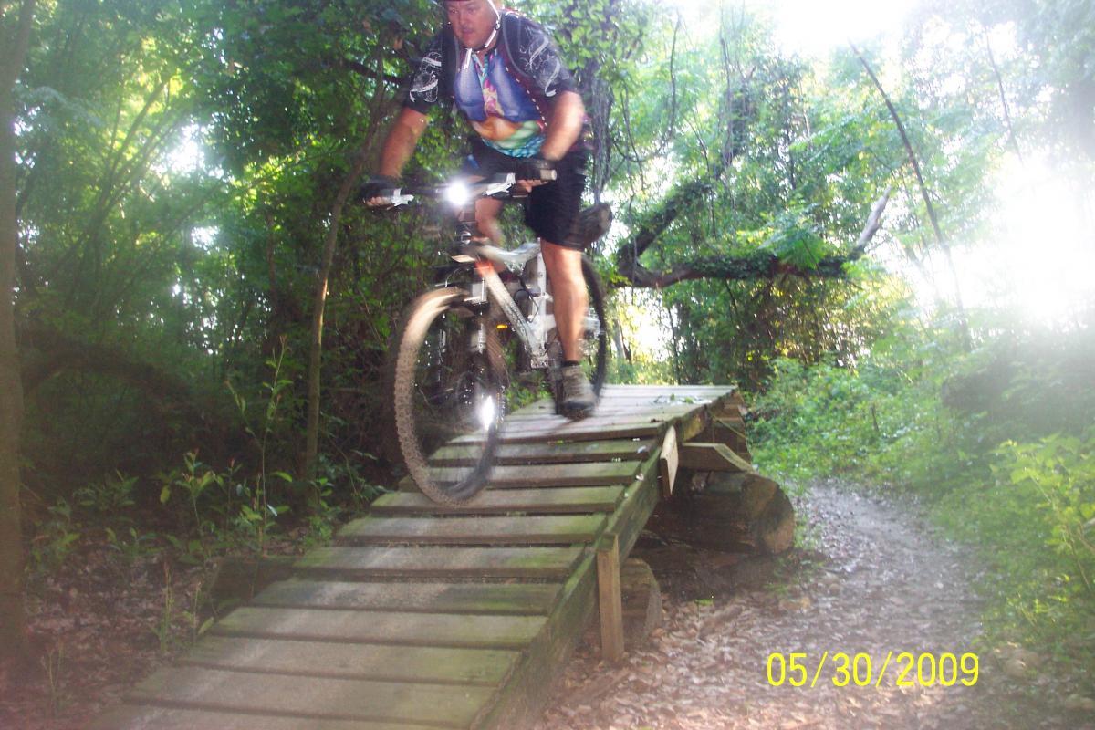 A mountain biker riding over a wooden bridge on a trail surrounded by lush green foliage and trees. The biker is captured mid-action, with dirt on the wheels and a focused expression, navigating the outdoor terrain. Loyce E. Harpe Park mountain bike trail.