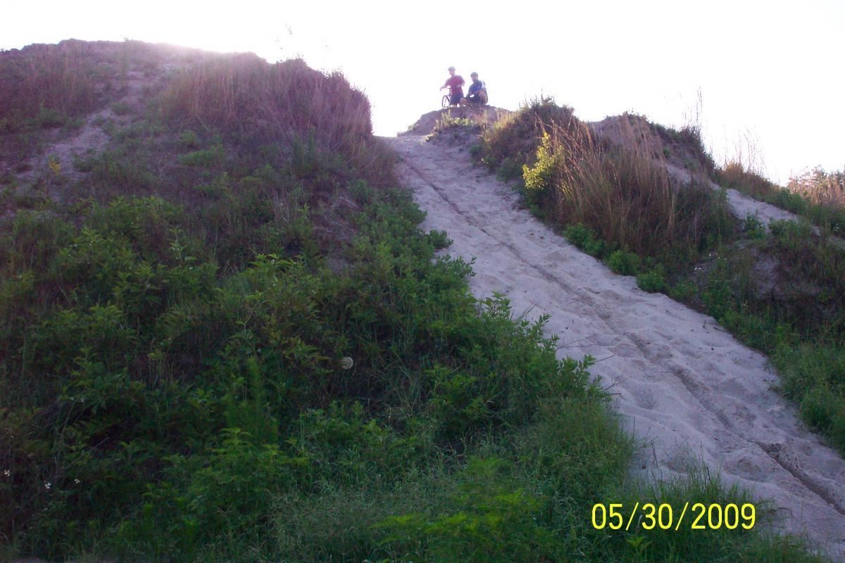 Two people sitting on a sandy hilltop surrounded by greenery, with a sloped path leading down. The sun is shining brightly in the background, creating a warm and sunny atmosphere. The image includes a date stamp at the bottom. Loyce E. Harpe Park mountain bike trail.