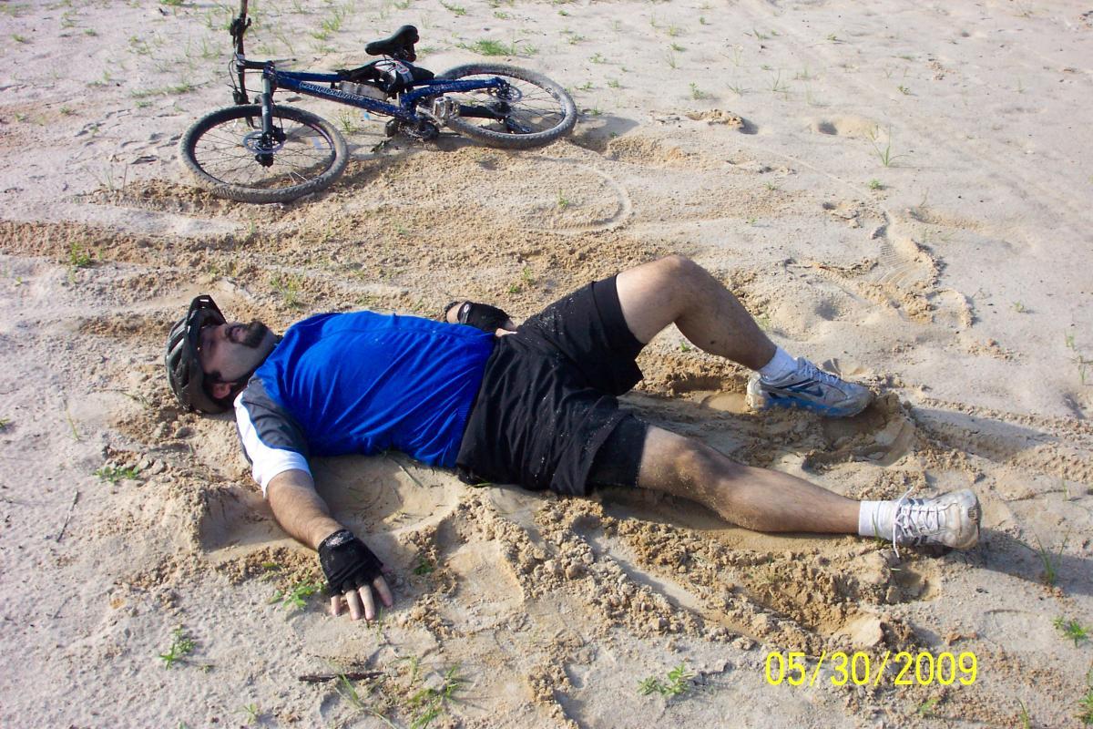 A person in athletic gear is lying on their back in sandy terrain, appearing tired or resting. A blue mountain bike is positioned nearby on the sand. The scene conveys a casual outdoor biking experience. Loyce E. Harpe Park mountain bike trail.