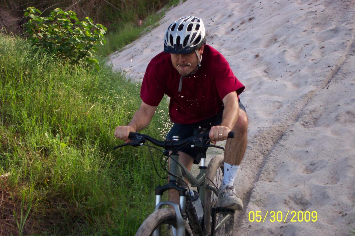 A person riding a mountain bike on a sandy trail, wearing a helmet and a red shirt, with dirt and water splashes visible on their clothing. Green grass and foliage are in the background, indicating an outdoor setting. Loyce E. Harpe Park mountain bike trail.