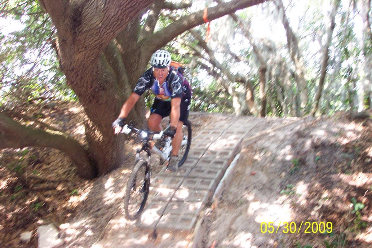 A mountain biker riding on a narrow wooden trail surrounded by trees, using a helmet and protective gear. The area appears wooded and somewhat rugged, suggesting an off-road biking location. The image is taken during daylight, with visible sunlight filtering through the leaves. Loyce E. Harpe Park mountain bike trail.