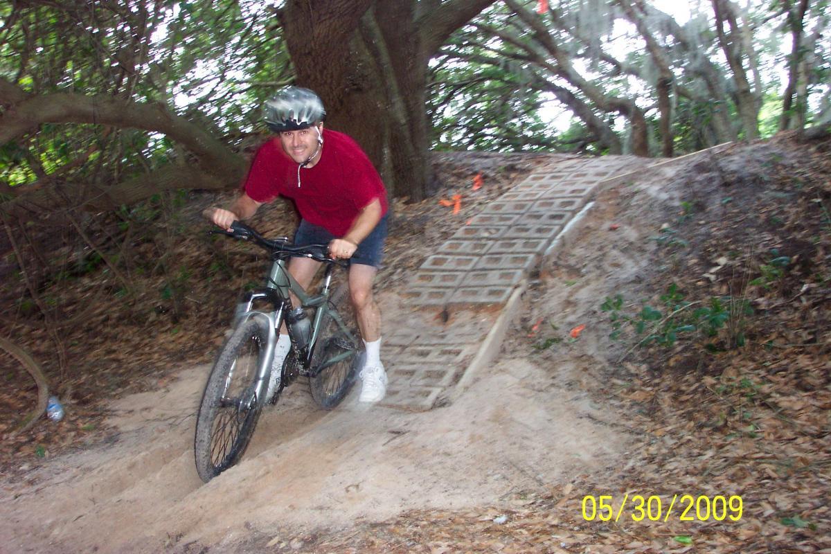 A person riding a mountain bike on a dirt track with a wooden ramp in a forested area. The cyclist is wearing a helmet and a red shirt, appearing to gain speed as they navigate the terrain. Trees and vegetation surround the path, and there are orange markers visible in the background. Loyce E. Harpe Park mountain bike trail.
