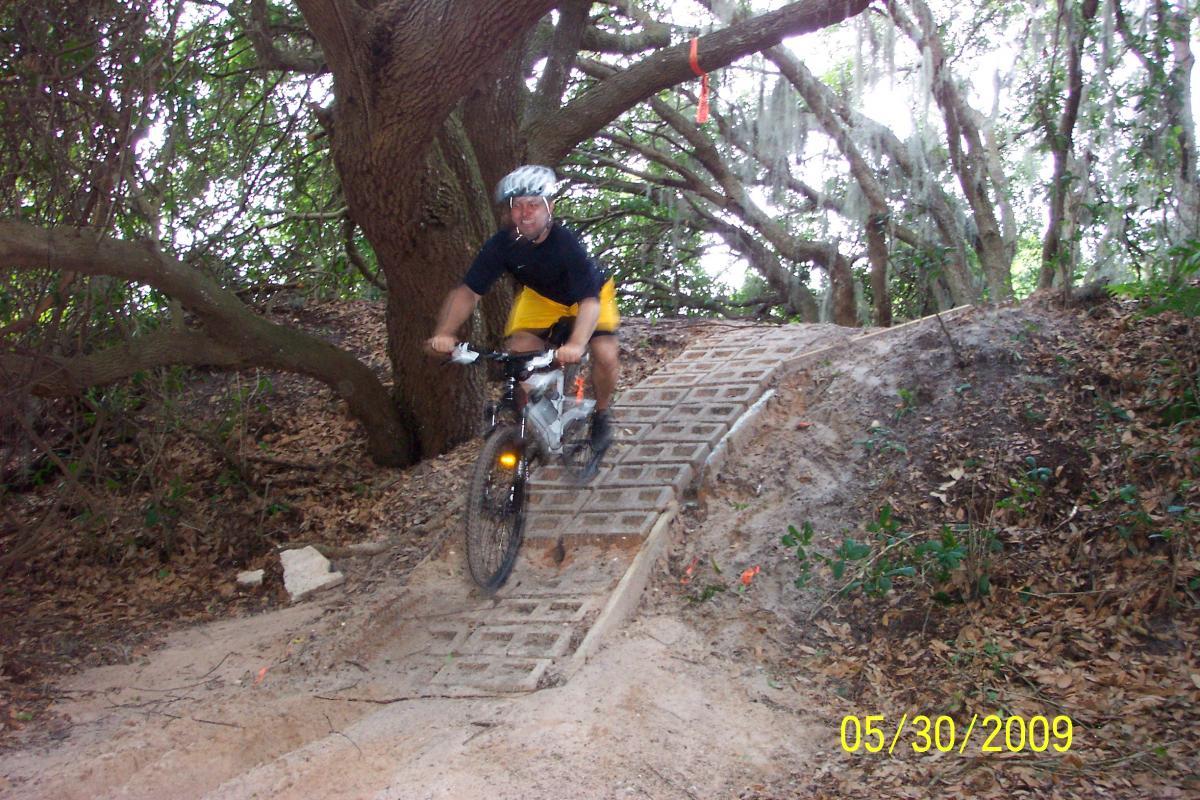 A mountain biker navigating a wooden ramp on a trail surrounded by trees and foliage. The rider is wearing a helmet and a black and yellow outfit, with the bike's front wheel slightly lifted as they descend the ramp. The scene captures the natural environment and the thrill of outdoor biking. Loyce E. Harpe Park mountain bike trail.