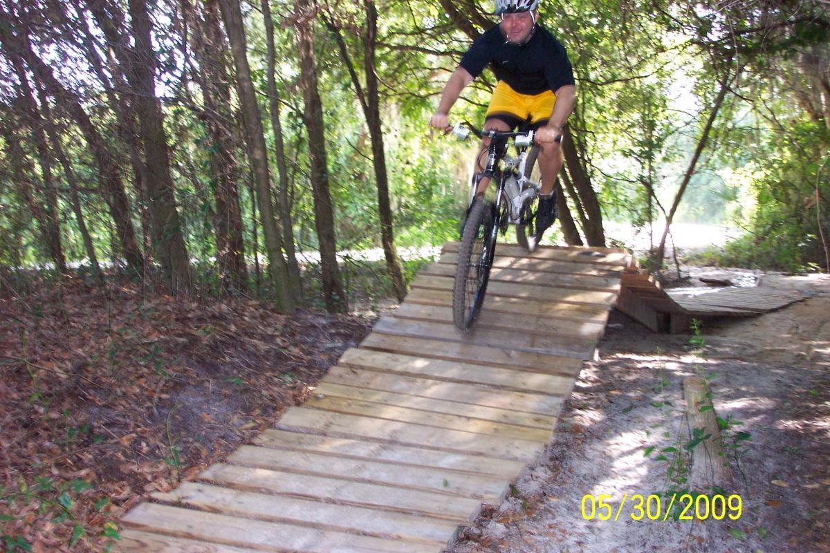 A cyclist in a black shirt and yellow shorts rides down a wooden ramp in a wooded area. The cyclist is in motion, navigating the ramp with trees and foliage in the background. Loyce E. Harpe Park mountain bike trail.