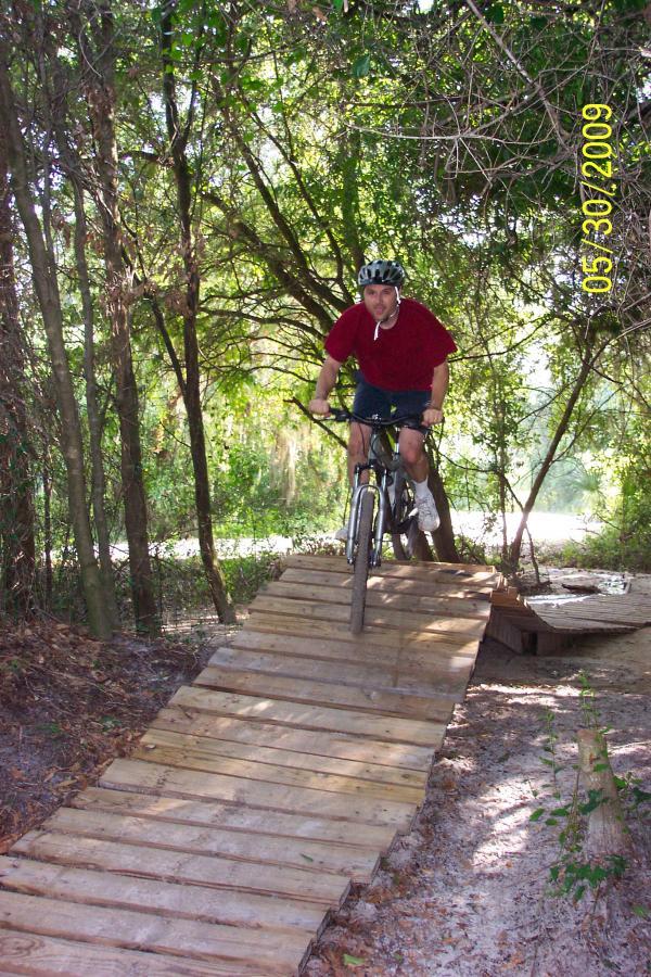 A person riding a mountain bike down a wooden ramp in a wooded area, surrounded by trees and greenery. The rider is wearing a helmet and a red shirt, demonstrating an action shot as they navigate the trail. Loyce E. Harpe Park mountain bike trail.