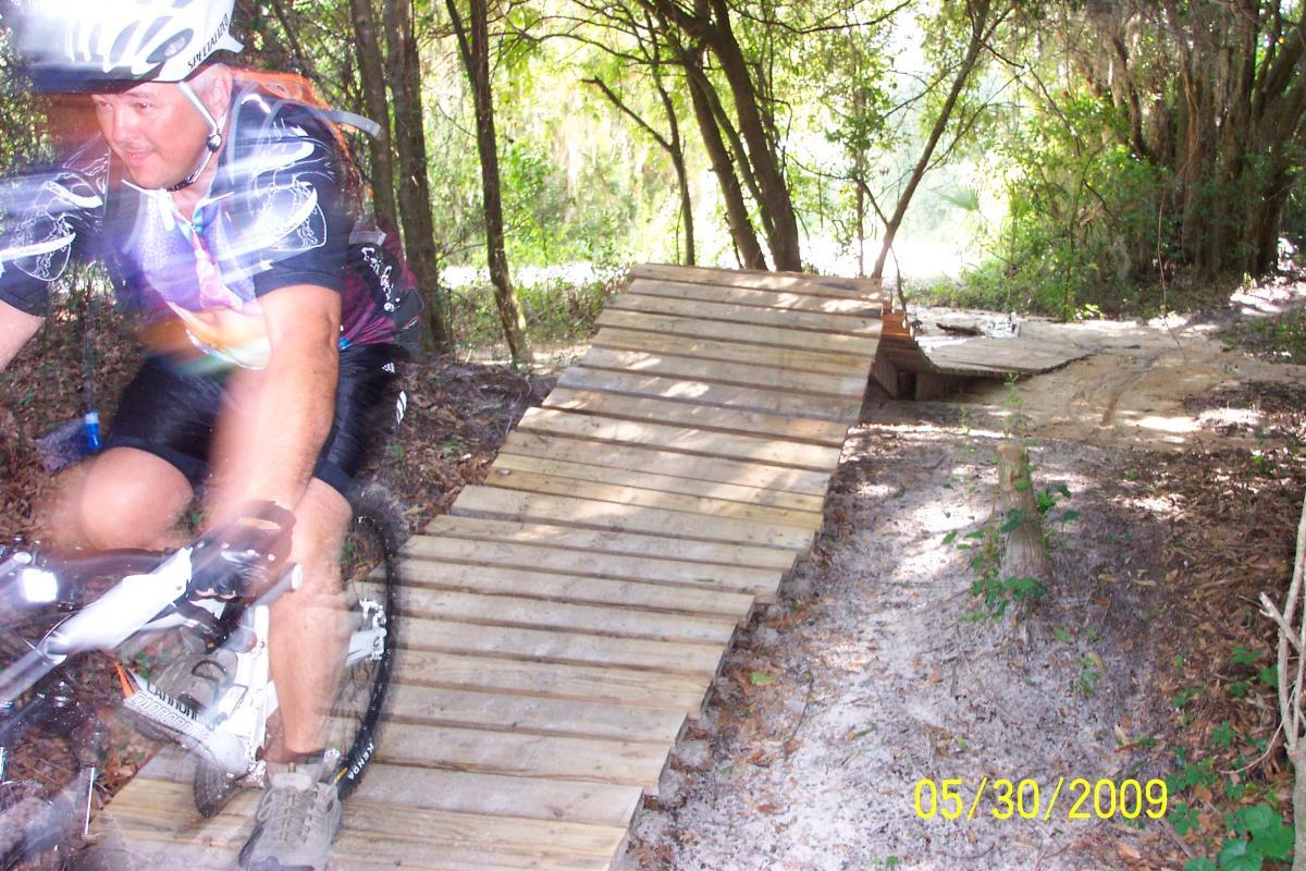 A mountain biker rides quickly over a wooden ramp on a trail surrounded by trees, with sandy ground and foliage nearby. The image captures the dynamic motion of the rider as he navigates the obstacle, showcasing the activity of mountain biking in a natural setting. Loyce E. Harpe Park mountain bike trail.