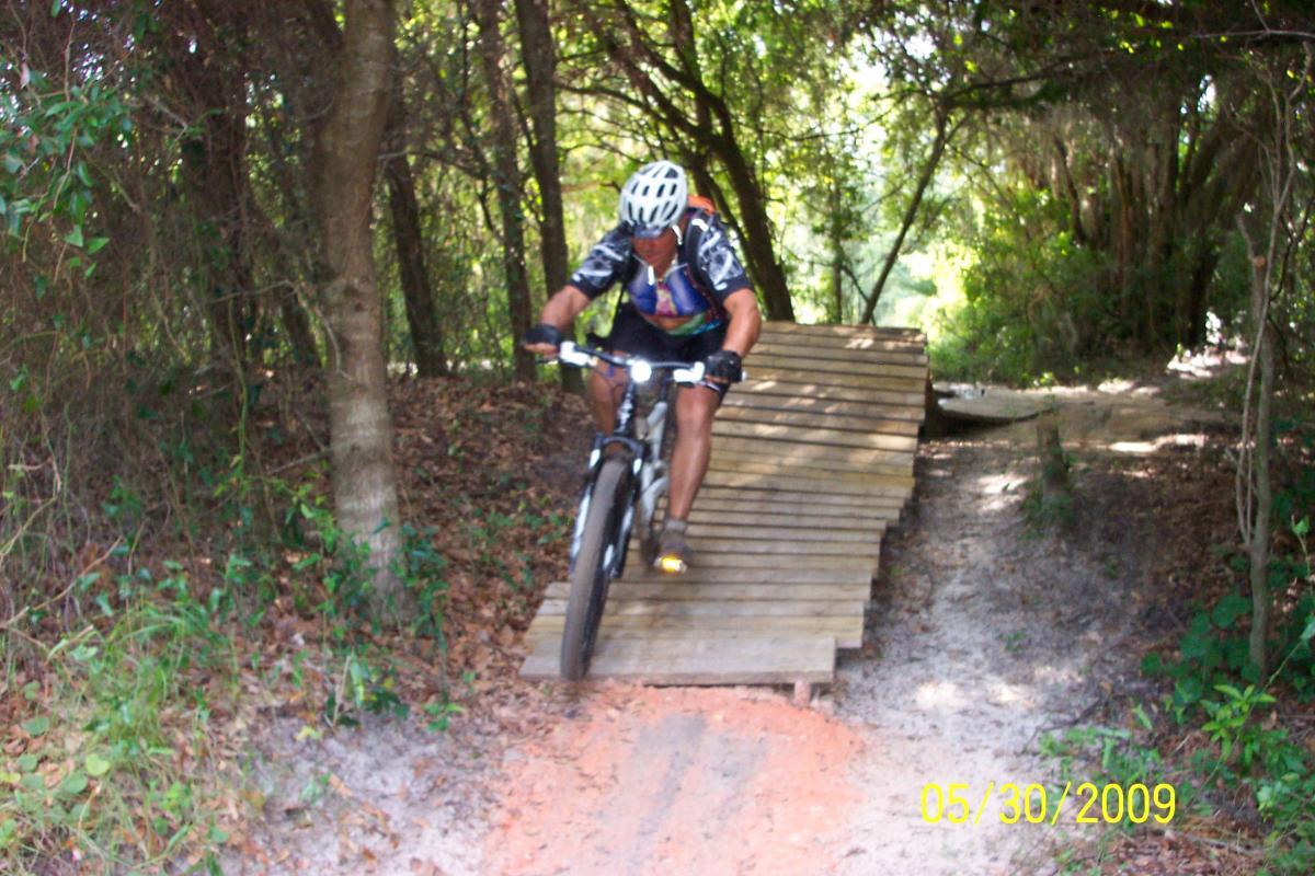A mountain biker descends a wooden ramp on a dirt trail surrounded by trees and greenery. The cyclist is wearing a helmet and cycling gear, focused on navigating the ramp. The scene captures an adventurous moment in an outdoor setting. Loyce E. Harpe Park mountain bike trail.