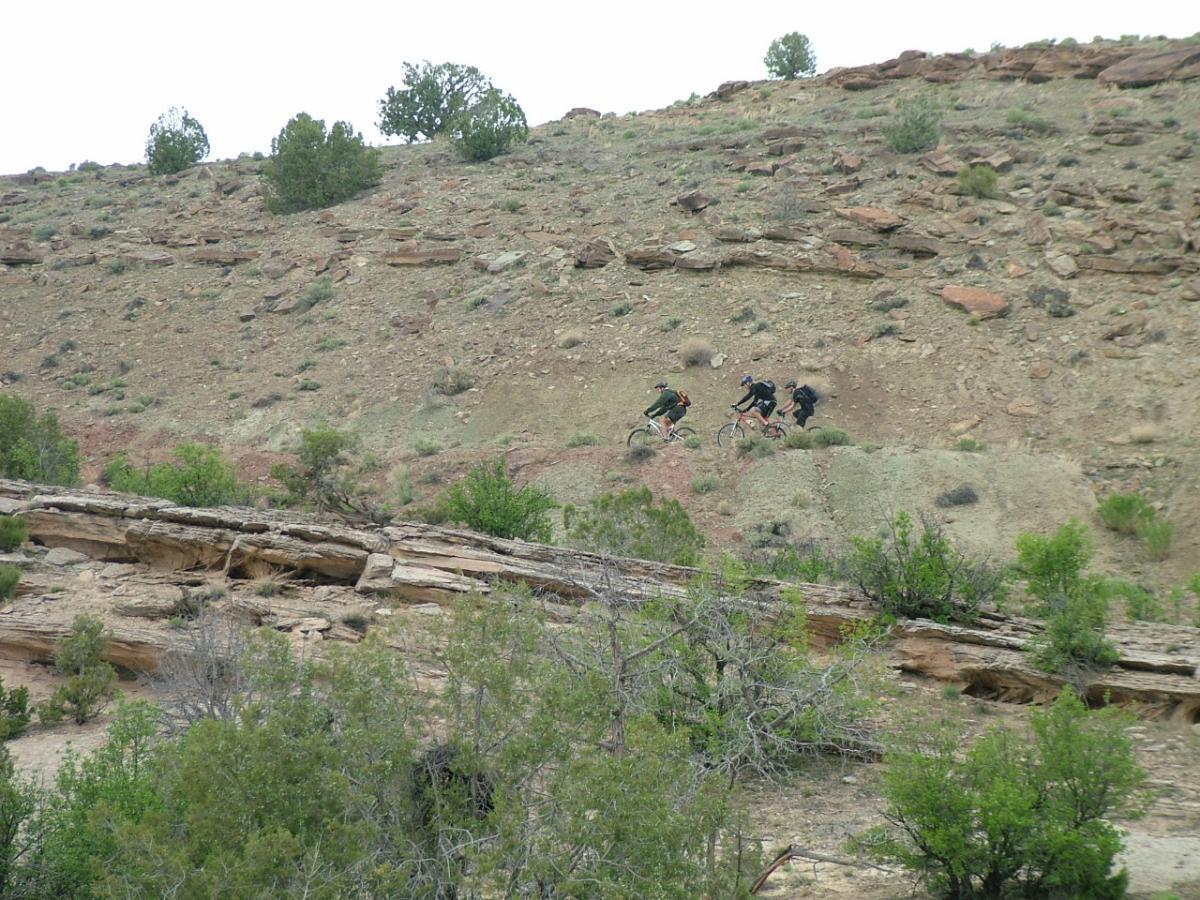 Two mountain bikers ride along a rugged, rocky hillside, surrounded by sparse vegetation and small trees. The terrain is dry and slightly sloped, with layers of exposed rock and soil visible. The sky is overcast, suggesting a cloudy day. Kokopelli Trail mountain bike trail.