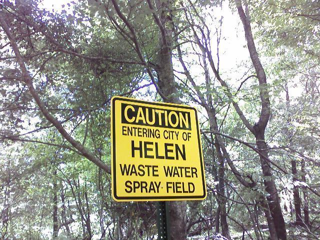 Yellow caution sign indicating "Entering City of Helen" with additional warning about "Waste Water Spray Field," surrounded by trees and foliage. Unicoi State Park mountain bike trail.