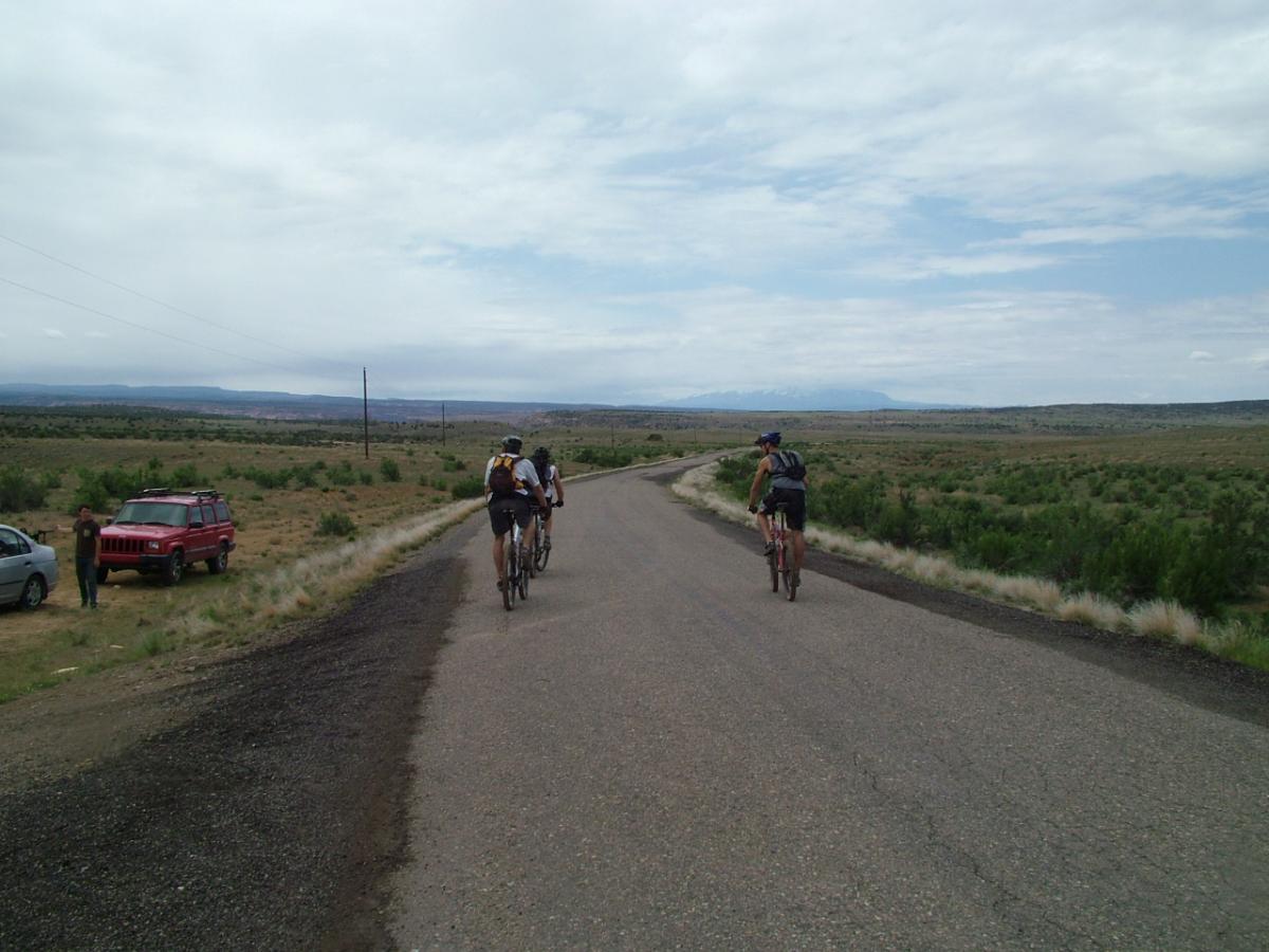 Three cyclists riding on a rural road, with a red SUV parked on the side. The landscape features open fields and distant mountains under a cloudy sky. Kokopelli Trail mountain bike trail.