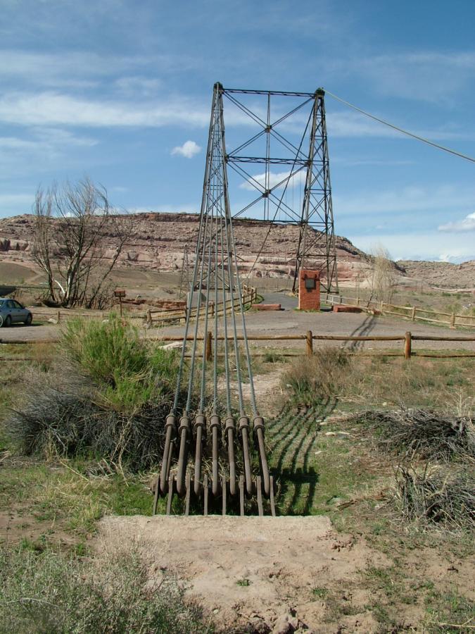A metal cable tower stands in a barren landscape, with several cables extending downward. In the background, there are rocky hills and a blue sky with scattered clouds. A small building and a parked car can be seen nearby, surrounded by sparse vegetation. Kokopelli Trail mountain bike trail.