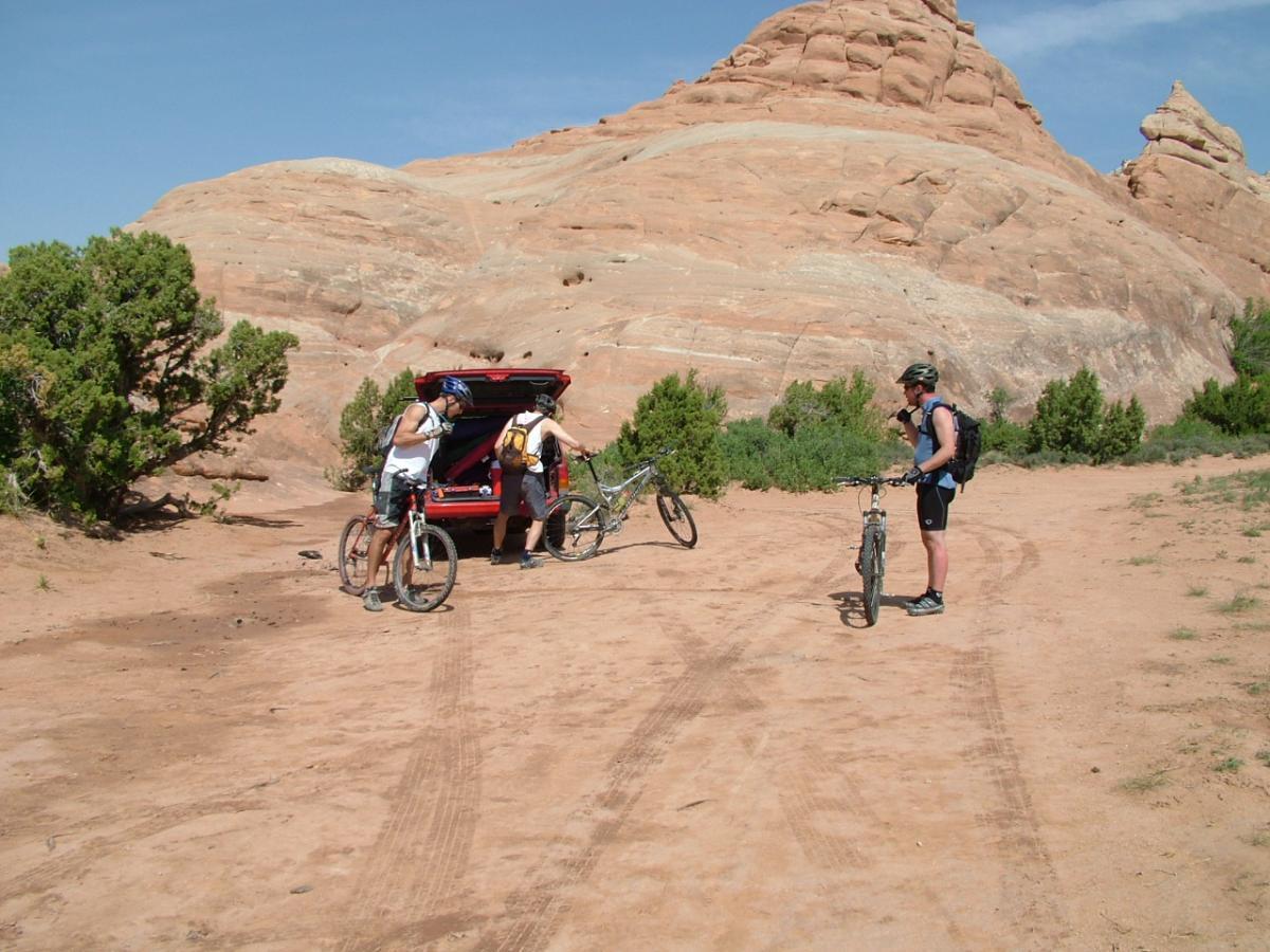 Three mountain bikers prepare for a ride in a desert landscape, with red rock formations in the background. One biker is at the back of a car, possibly retrieving gear, while another adjusts their bike. The scene is sunny with a clear blue sky, and there are shrubs and tire tracks in the sandy terrain. Kokopelli Trail mountain bike trail.