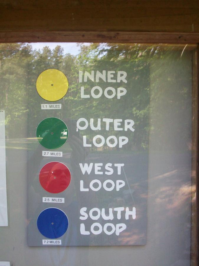 Sign displaying trail loop information. It includes four sections: "INNER LOOP" marked with a yellow circle and measuring 1.1 miles, "OUTER LOOP" marked with a green circle at 2.7 miles, "WEST LOOP" with a red circle at 2.5 miles, and "SOUTH LOOP" marked with a blue circle at 7.2 miles. The background shows trees and natural surroundings. Beaver Dam mountain bike trail.