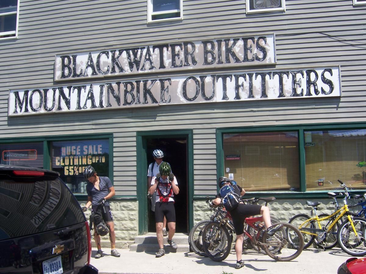 Image of the exterior of Blackwater Bikes, a mountain biking outfitter. The building features a sign that reads "BLACKWATER BIKES MOUNTAINBIKE OUTFITTERS." In front of the store, two cyclists are adjusting their gear, while a third cyclist is entering. Several bicycles are parked nearby, and a sign in the window advertises a sale on clothing. The scene is set on a sunny day with clear visibility.