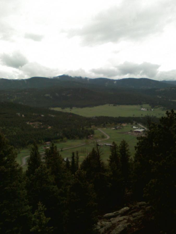 A scenic view from a mountain overlooking a green valley surrounded by forests and distant mountains under a cloudy sky. There are patches of farmland and a winding road visible in the valley. 3 Sisters / Alderfer mountain bike trail.