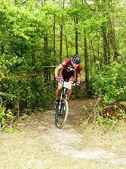 A mountain biker in a red jersey and helmet navigating a dirt trail surrounded by greenery in a forested area. Cypresswood Trails mountain bike trail.