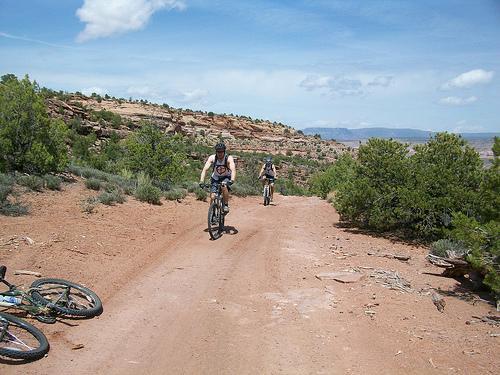 Two mountain bikers ride on a dirt trail surrounded by desert vegetation and rocky terrain under a blue sky with scattered clouds. In the foreground, another bike lies on the ground. Kokopelli Trail mountain bike trail.