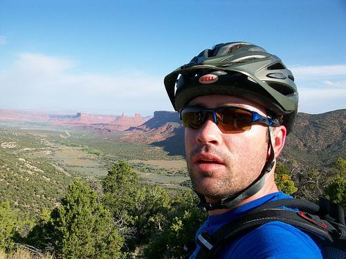 A person wearing a helmet and sunglasses poses for a selfie while mountain biking, with a scenic view of red rock formations and lush green valleys in the background. Kokopelli Trail mountain bike trail.