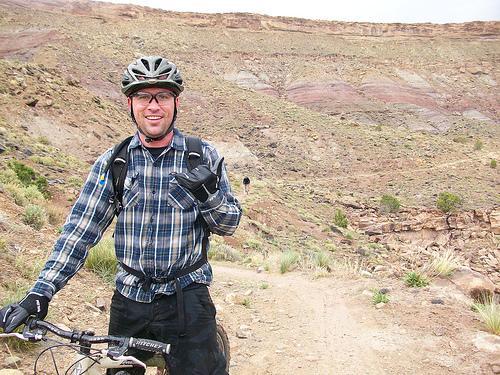 A man wearing a helmet and a plaid shirt is standing next to a mountain bike on a dirt trail. He is smiling and giving a thumbs-up gesture, with a rocky landscape featuring layered hills in the background. The scene appears to be in a remote outdoor area suitable for biking. Kokopelli Trail mountain bike trail.