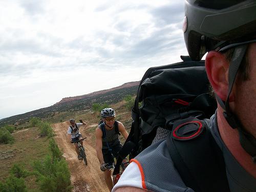 A group of mountain bikers on a dirt trail surrounded by greenery and rocky terrain, with one cyclist in the foreground and another in the background. The riders are wearing helmets and carrying backpacks, indicating an outdoor adventure. The sky is partly cloudy, suggesting a mild day for biking. Kokopelli Trail mountain bike trail.