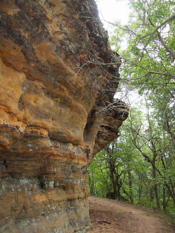 A close-up view of a rugged rock formation, featuring textured layers of orange and brown stone, alongside a narrow, winding dirt path. Lush greenery, including trees and foliage, frames the scene, highlighting the natural landscape. Levis Mounds mountain bike trail.