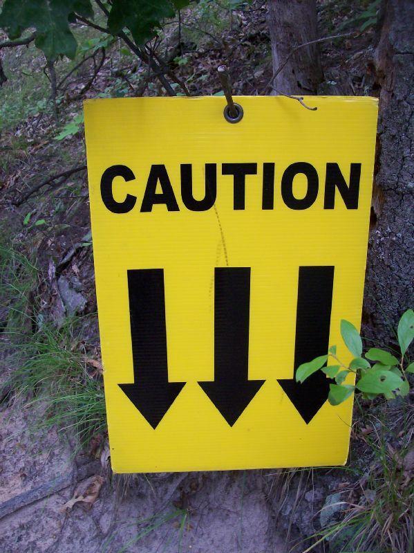 A bright yellow caution sign with bold black lettering that reads "CAUTION" at the top, featuring three downward-pointing black arrows below. The sign is positioned near trees and grass in a natural outdoor setting. Levis Mounds mountain bike trail.