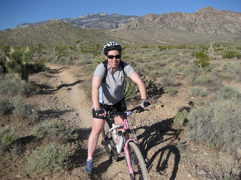 A woman wearing a helmet and sunglasses is riding a mountain bike along a dirt trail in a desert landscape. She is smiling and appears to be enjoying the outdoor adventure, with mountains in the background and sparse vegetation surrounding the pathway. Blue Diamond mountain bike trail.