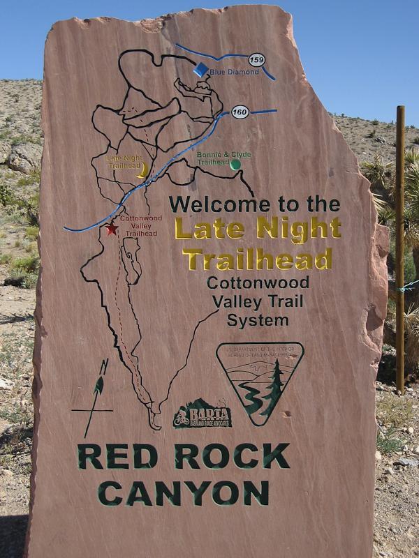 A stone sign indicating the "Late Night Trailhead" for the Cottonwood Valley Trail System in Red Rock Canyon. The sign features a map with marked trails, including the Cottonwood Valley Trailhead and the Bonnie & Clyde Trailhead. Directions and details about the trail system are displayed in yellow and green text on a reddish-brown background. Blue Diamond mountain bike trail.